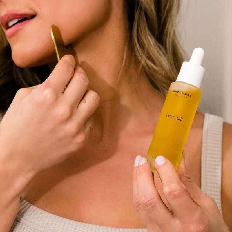 Hydrating Face Oil bottle next to stainless steel Gua Sha tool on white background
